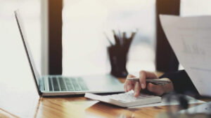 A person doing their taxes while using a white calculator and laptop.