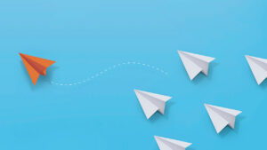 An orange paper airplane flying away from a group of white paper airplanes.